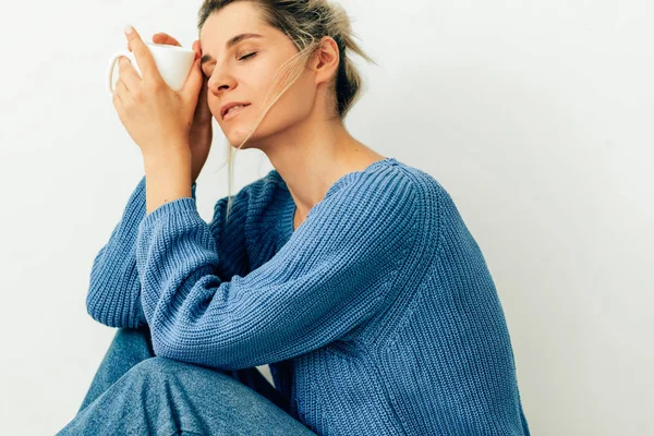 Sevimli genç kadın elinde kapuçino bir kupa ile rahat örme mavi kazak ve kot giyiyor. Güzel genç kadın evde sıcak içecek içiyor. Bir kahve fincanı tutan lovely sarışın kadın 