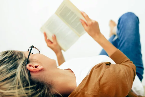 Afbeelding van de jonge vrouw lezen boek, het dragen van transparante brillen liggend op het tapijt in hygge huis. Vrouwelijke ontspannen in een gezellige woonkamer, het lezen van een boek. Vrouwelijke student zittend thuis, studeren. — Stockfoto