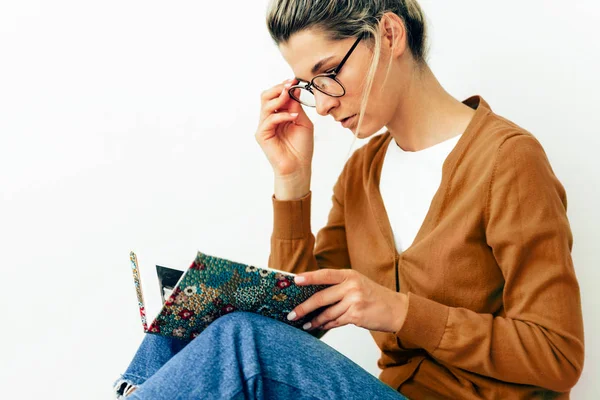 Bela jovem mulher lendo livro, vestindo óculos transparentes sentados no tapete em casa hygge. Feminino relaxante na acolhedora sala de estar, lendo um livro. Estudante do sexo feminino sentado em casa, estudando . — Fotografia de Stock