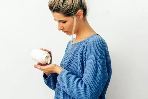 Side view image of woman's hand pours the pills out of the bottle. Young blonde female wearing blue knitted sweater pouring some pills in her hand from a white bottle. Healthcare and medicine. — Stock Photo, Image