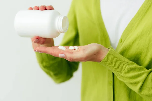 Cropped studio image of woman\'s hand pours the pills out of the bottle. Healthy female wearing green cardigan and white t shirt pouring some pills in her hand from white bottle. Healthcare, medicine