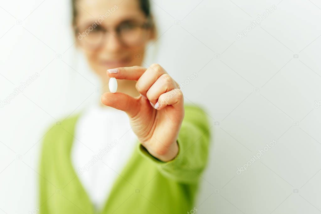 Image of focused pill in hand with blurred view of smiling woman. Beautiful young woman wearing green cardigan and white t-shirt taking pill isolated on white wall. Vitamin, dietary Supplements.