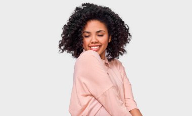 Pembe shrit giymiş koyu tenli genç kadın memnun, mutlu hissediyor. Afro-Amerikan kadın beyaz duvar üzerinde poz, geniş gülümseyen