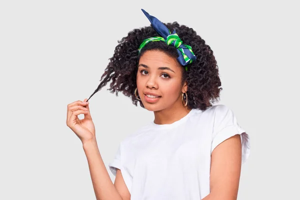 Dunkelhäutige junge Frau in weißem T-Shirt und Stirnband, spielt mit den Haaren, fühlt sich glücklich. Afrikanische Amerikanerin lächelt und posiert im Studio. — Stockfoto