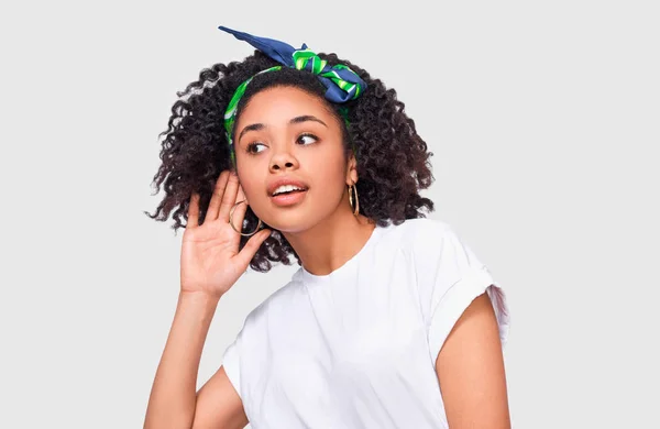 Jovem afro-americana prestando atenção e colocando a mão na orelha pedindo a alguém para falar mais alto ou sussurrar, isolado na parede branca. Menina afro bonita que ouve conversa secreta — Fotografia de Stock