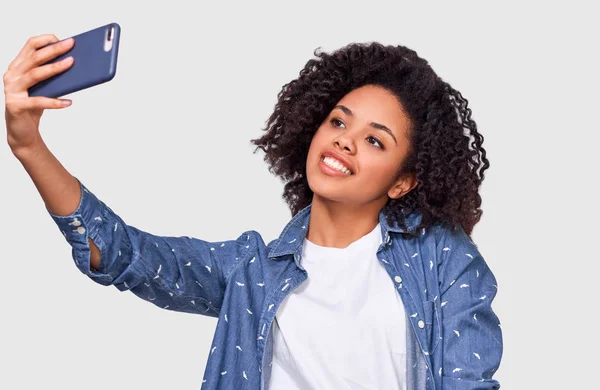 Gorgeous mörkhyade ung kvinna i vit t-shirt och blå skjorta, tar Selfie över vit Studio vägg. Lycklig afrikansk amerikansk kvinnlig leende, tar en selfie på hennes smartphone. — Stockfoto