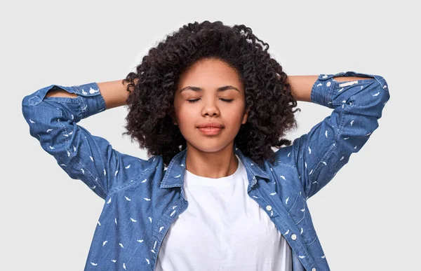 Die verträumte dunkelhäutige junge Frau in Jeanshemd und weißem T-Shirt, die Hände auf dem Kopf, fühlt sich glücklich. Afroamerikanerin entspannt mit geschlossenen Augen vor weißer Wand posierend — Stockfoto