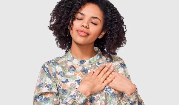 La imagen franca de la joven afroamericana mantiene ambas palmas en el pecho con los ojos cerrados, vestidas con camisa floral, aisladas sobre fondo blanco. Personas, emociones, salud, concepto de lenguaje corporal —  Fotos de Stock