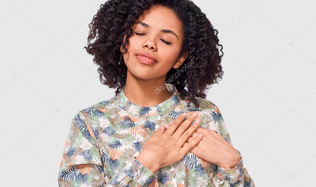 Candid image of African American young woman keeps both palms on the chest with closed eyes, dressed in floral shirt, isolated over white background. People, emotions, health, body language concept