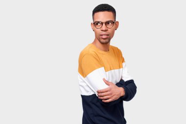 Renkli kazak giyen çekici Afro-Amerikan genç adam, beyaz stüdyo arka plan üzerinde gözlük ile bir tarafa bakarak, çapraz eller tutar. Stüdyoda poz veren kendine güvenen erkek öğrenci