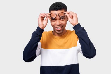 Gülümseyen genç Afro-Amerikan adam giyen ve yuvarlak trendy şeffaf gözlük ile bakıyor. Afro öğrenci erkek beyaz stüdyo duvarında poz renkli kazak giyiyor. İnsan kavramı 