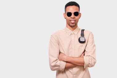 Omuzlarında kulaklık olan Afro-Amerikan genç adamın portresi kameraya bakıyor. Afro erkek öğrenci güneş gözlüğü giyiyor, rahat gömlek, beyaz stüdyo duvarında poz