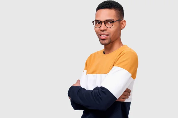 Portrait horizontal en studio d'un jeune homme afro-américain agréable portant un pull à la mode garde les mains croisées, regardant la caméra à travers des lunettes transparentes. heureux mâle étudiant posant dans studio . — Photo