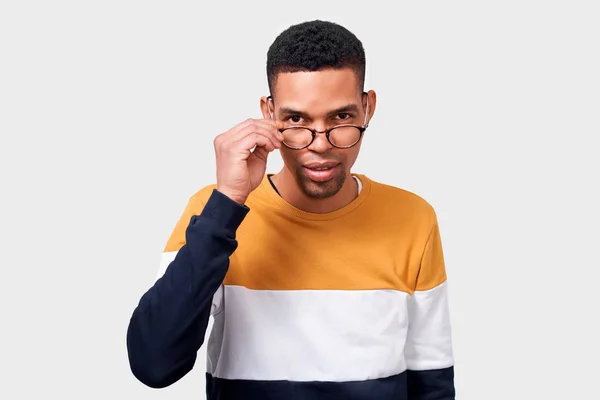 Porträt eines ernsthaften jungen afrikanisch-amerikanischen Mannes, der eine runde trendige durchsichtige Brille trägt und durch sie blickt. afro student male in bunten pullover posiert auf weißen studio-wand. Menschen-Konzept — Stockfoto