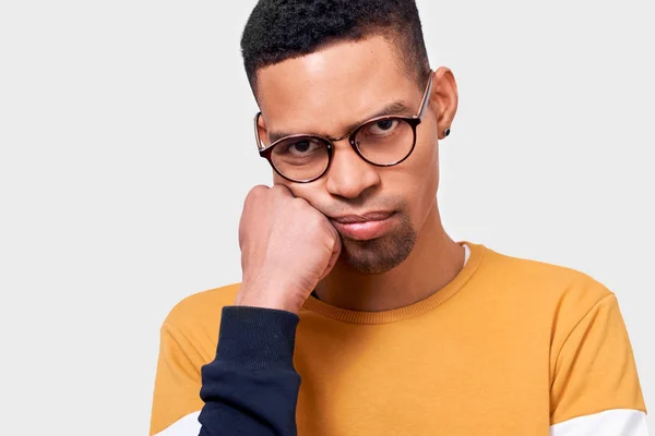 Primer plano retrato de hombre afro cansado se apoya en la mano, tiene expresión disgustado, vistiendo suéter y gafas, posando en el estudio. Un afroamericano sobrecargado de trabajo se siente aburrido. Personas, sentimientos, emociones — Foto de Stock