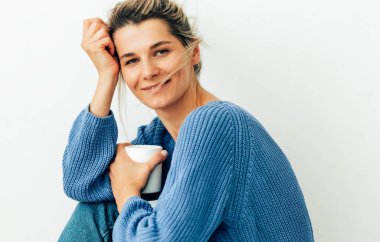 Sevimli genç kadın kameraya bakarak çay beyaz bir fincan ile rahat örme mavi kazak giymiş. Güzel genç kadın evde sıcak içecek içiyor. Sarışın kadın bir kahve fincanı tutan. 