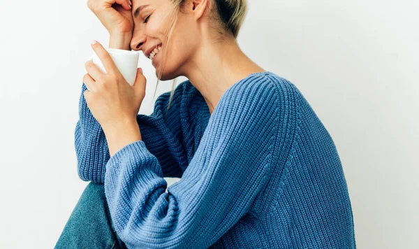 Feliz linda jovem vestida com camisola azul de malha aconchegante com uma caneca branca com um chá na mão. Mulher bonita bebendo bebida quente em casa. Sorrindo loira fêmea segurando uma xícara de café . — Fotografia de Stock