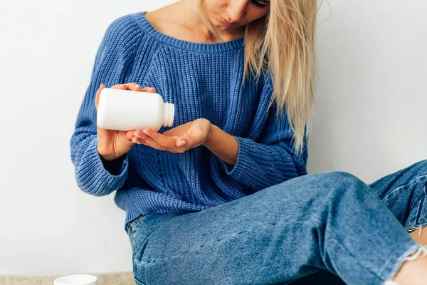 Bijgesneden beeld van jonge vrouw giet de pillen uit de fles. Jonge blonde vrouw dragen van blauwe gebreide trui gieten sommige pillen in haar hand van een witte fles. Gezondheidszorg en geneeskunde. — Stockfoto