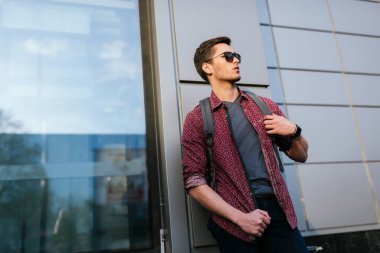 Moda güneş gözlüğü ile Casual yakışıklı kafkas adam, sokak şehir uzağa bakıyor. Çekici işadamı ve turist sırt çantası ile şehirde açık havada yürüyüş. Kentsel yaşam tarzı kavramı.