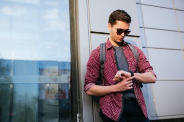 Moda güneş gözlüğü ile Akıllı genç yakışıklı kafkas adam, saat bakarak, sokak şehirde. Çekici işadamı ve turist sırt çantası ile şehirde açık havada yürüyüş. Kentsel yaşam tarzı kavramı.