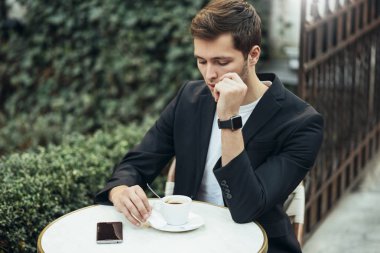Beyaz ciddi ofis çalışanı, öğle tatili sırasında mesafeli çalışma için tablet kullanarak resmi takım elbise giyiyor, bir kafede masada oturan, şehirde bir fincan kahve içen. 