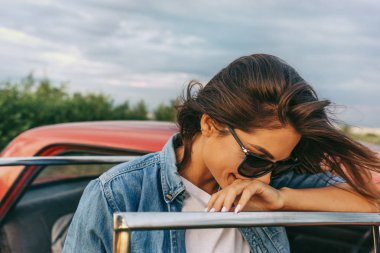 Genç beyaz bir kadın bulutlu bir zeminde kırmızı arabasının yanında güneş gözlüğü takıyor. Bir alan arka plan üzerinde saç üfleme ile Avrupa kadın açık hava gezisi. Seyahat konsepti.