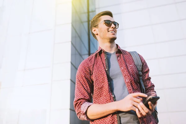 Концепция городской моды. Нижний вид на счастливый веселый привлекательный кавказский мужчина в стильных солнечных очках в городе. Молодой бизнесмен, использующий смартфон на улице. Путешественник . — стоковое фото