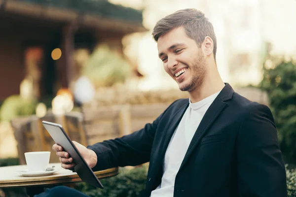 Horizontal, happy caucasian man look to the digital tablet sitting in cafe with cup of coffee. Business man having breakfast sitting on beautiful terrace with plants. Reading and browsing internet. — Stock Photo, Image