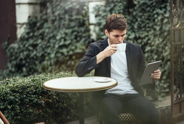 Chytrý fešák běloch kontroluje jeho zařízení a pije horkou kávu. Mladý veselý chlapík v obleku si užívá ráno a čte zprávy o dni v kavárně na terase venku ve městě — Stock fotografie