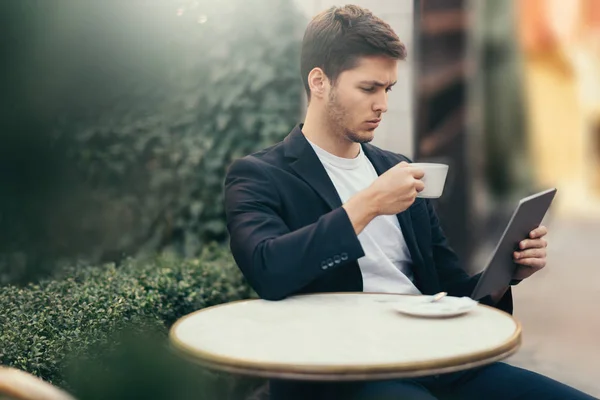 Yakışıklı ciddi kafkas adam kahve içmek ve kafede oturan dijital tablet bakmak. Bitkilerle güzel bir terasta oturarak kahvaltı alan iş adamı. Okuma ve internet tegezİş. — Stok fotoğraf