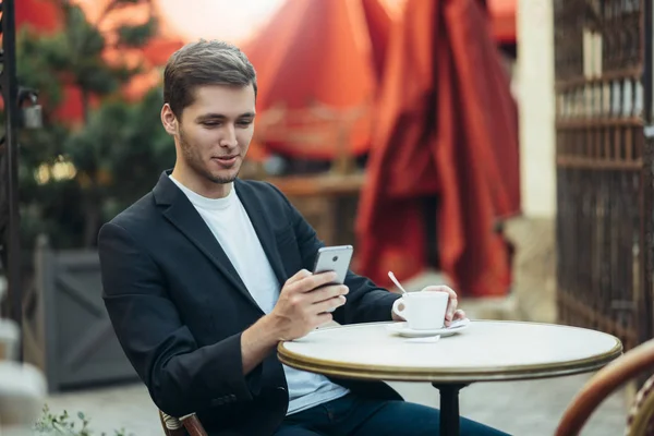 Happy stylish Caucasian businessman in suit drinking cappccino at coffee shop, sitting on terrace with cell phone. Business, modern technology, communication and people concept. — Stock Photo, Image