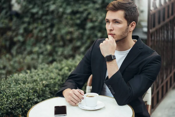 Portait of formal Caucasian serious office worker wearing formal suit thinking about new project, sitting at the table at a cafe, having a cup of coffee in the city.