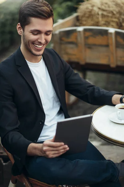 Pohledný muž s úsměvem v příležitostné práci kontroluje e-maily nebo čte Světové novinky o digitální tabletové kavárně, která sedí u stolu v útulné kavárně. Koncepce připojení a technologie. — Stock fotografie