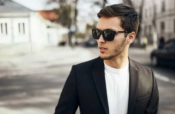 Urban fashion portrait of young handsome businessman in casual suit in dark sunglasses sitting in the street goind to the work and looking aside.