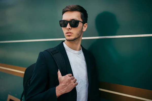 Stylish fashion young Caucasian man in sunglasses and black jacket with backpack walking over green city background. Fashionable business tourist travel around the world for new project and clients.