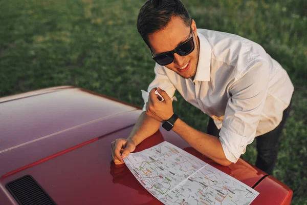 Handsome young male wears white shirt and sunglasses using a map on a road trip for guide destination. Tourist man looking away while standing next to hood of the car, lost on nature. Travel, people