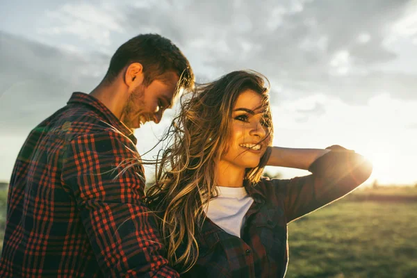 Gün batımını kucaklayan çayırda birlikte duran romantik çiftin çekici açık hava portresi. Aşk romantik yakışıklı adam doğa hayran onun güzel kız arkadaşı sarılma ekose gömlek giyiyor.