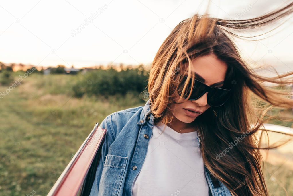 Beautiful attractive young Caucasian woman wearing sunglasses near to her red car on cloudy background. European female outdoor trip with blowing hair on a field background. Travel concept.