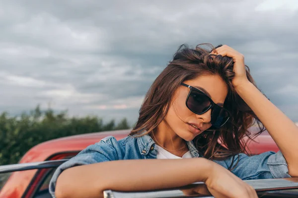 Romântico bonito atraente jovem mulher caucasiana usando óculos de sol perto de seu carro vermelho no fundo nublado. Viagem ao ar livre feminino europeu com cabelo soprando em um fundo de campo. Conceito de viagem . — Fotografia de Stock