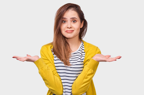 Questioned young woman, looking uncertain during discussing, dressed in striped t-shirt and yellow cardigan, posing over white wall. Student girl has confused expression. People emotions