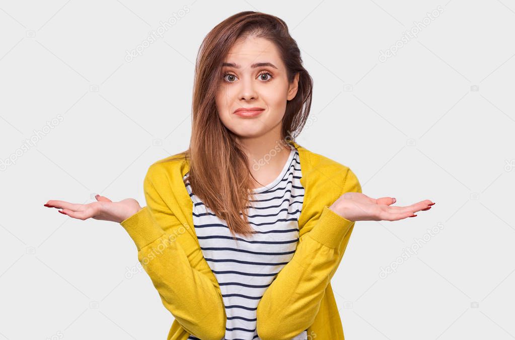 Questioned young woman, looking uncertain during discussing, dressed in striped t-shirt and yellow cardigan, posing over white wall. Student girl has confused expression. People emotions