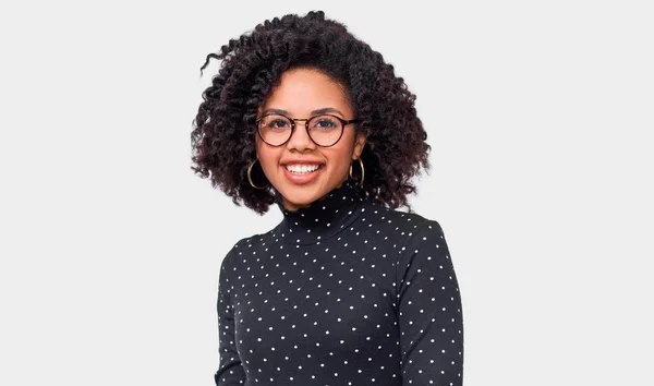 Retrato del estudio de la joven afroamericana vestida de negro con puntos blancos camisa de manga larga, sentirse feliz. Afro hembra sonriendo ampliamente, usando gafas redondas transparentes posando sobre pared blanca —  Fotos de Stock