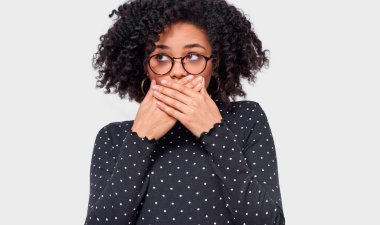Close-up portrait of horrified African American young woman covering mouth with hands feeling scared, looking aside with bulging eyes, posing over white wall. Afro female student with shocked face. clipart
