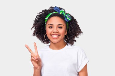 Büyüleyici Afro-Amerikan genç kadın geniş gülümseyerek, kameraya bakarken barış jesti gösteren, beyaz t-shirt ve yeşil kafa bandı giyen, beyaz stüdyo arka plan karşı duran. 
