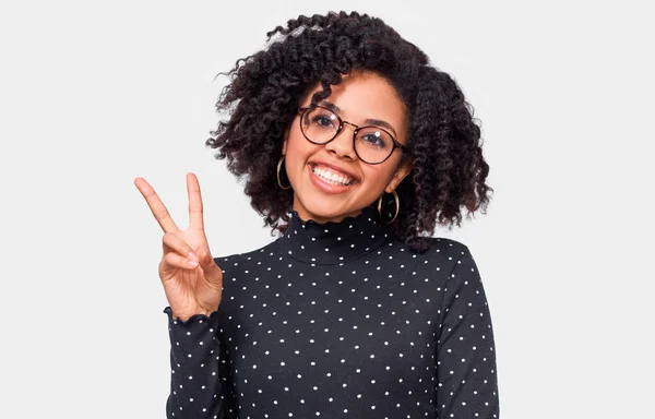 Mujer joven afroamericana positiva sonriendo ampliamente, mostrando gesto de paz mientras mira a la cámara, de pie sobre fondo blanco del estudio . —  Fotos de Stock