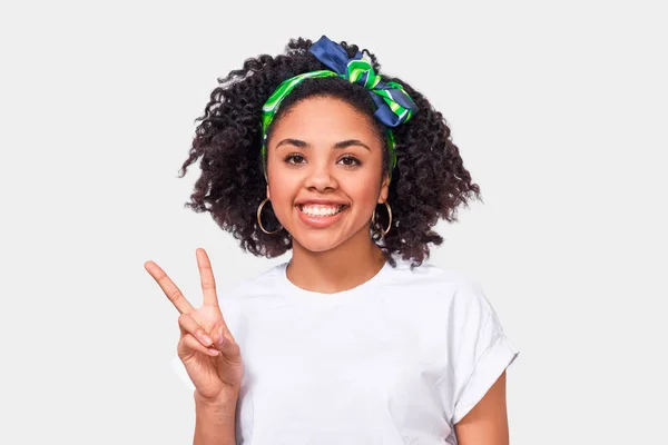 Charmante junge afrikanisch-amerikanische Frau lächelt breit, zeigt Friedensgeste, während sie in die Kamera blickt, trägt weißes T-Shirt und grünes Stirnband, steht vor weißem Studiohintergrund. — Stockfoto
