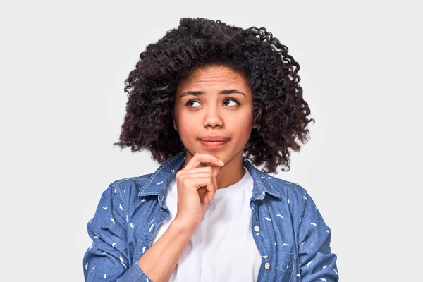 La dudosa mujer afroamericana tiene expresión desconcertante, frunce el ceño y mira a un lado con la mano en la barbilla. Mujer de piel oscura no puede elegir, ha cuestionado la expresión, aislado en blanco . —  Fotos de Stock