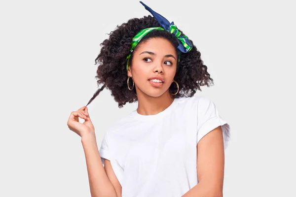 Retrato de una joven de piel oscura con camiseta blanca y diadema verde, jugando con un pedazo de su pelo rizado, soñando y sintiéndose feliz. Mujer afroamericana sonriendo y posando en el estudio . — Foto de Stock