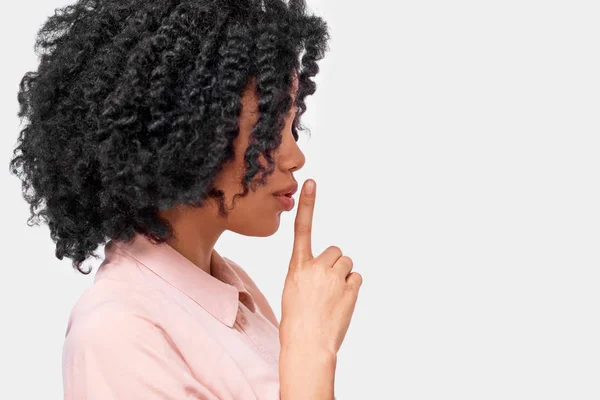 Retrato de vista lateral de una joven afroamericana que usa camisa rosa, sosteniendo el dedo índice en los labios, pidiendo guardar silencio. Mujer de piel oscura pide estar tranquilo . —  Fotos de Stock