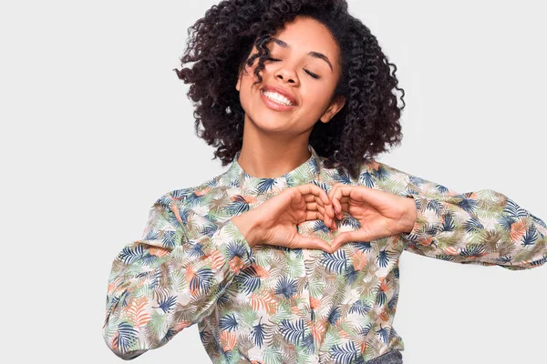 Souriante jeune femme afro-américaine en forme de cœur avec les doigts. Jolie chemise à motifs floraux habillée de femmes afro, montrant un geste de coeur avec les deux mains, signe d'amour, posant sur fond blanc — Photo